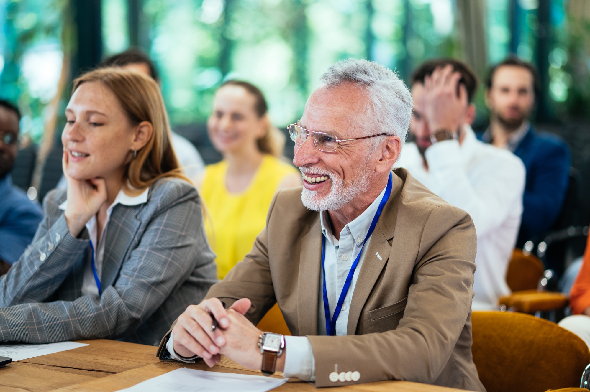 Smiling senior man in a beige suit and glasses attending a conference, seated next to a young woman in a gray blazer, with a blurred background of other participants.