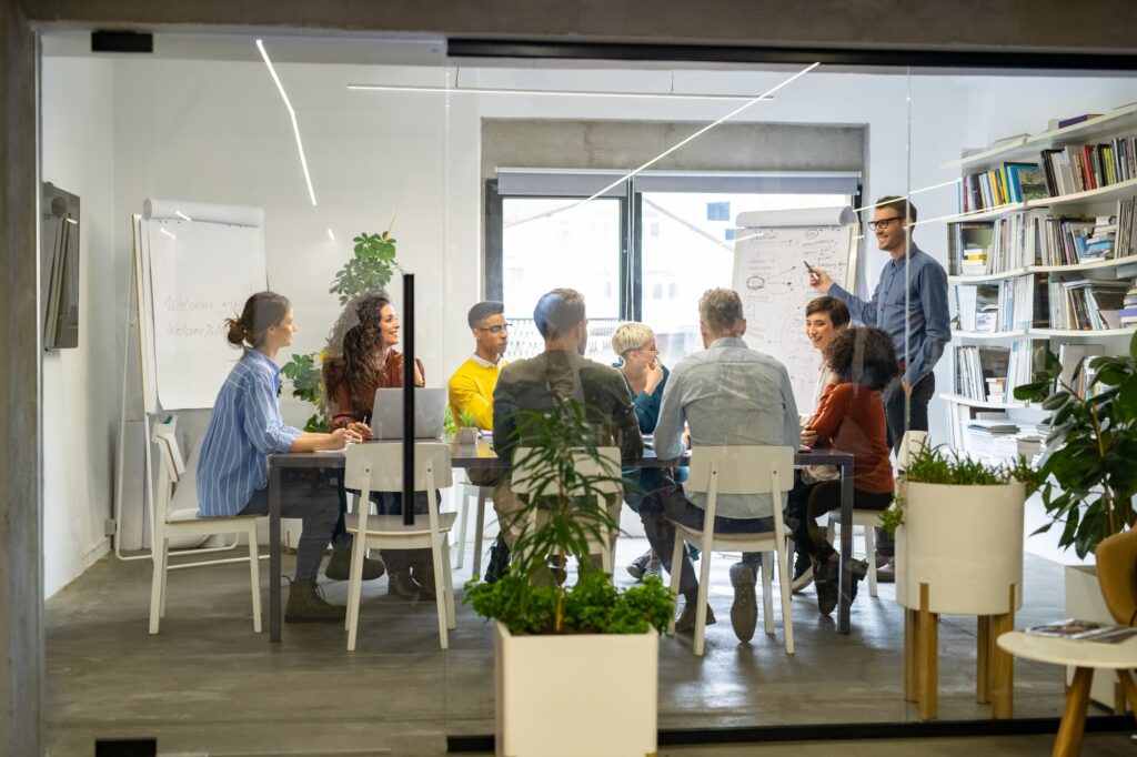 Team of diverse professionals in a modern office participating in a collaborative meeting, with a presenter pointing at a flipchart."